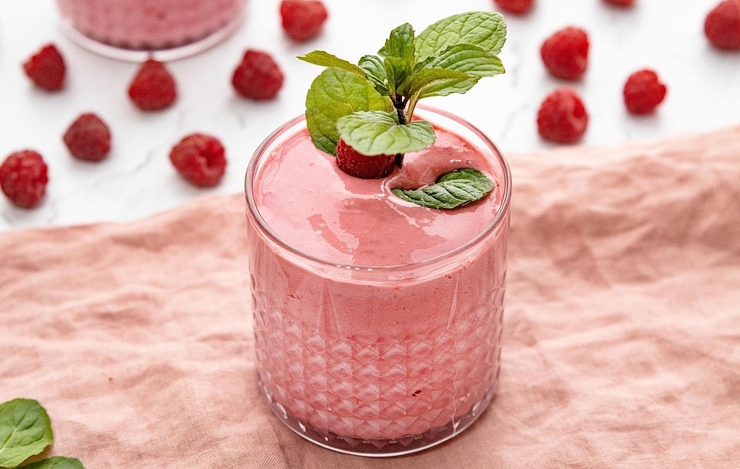 A pink smoothie in a small glass, with raspberries scattered around it