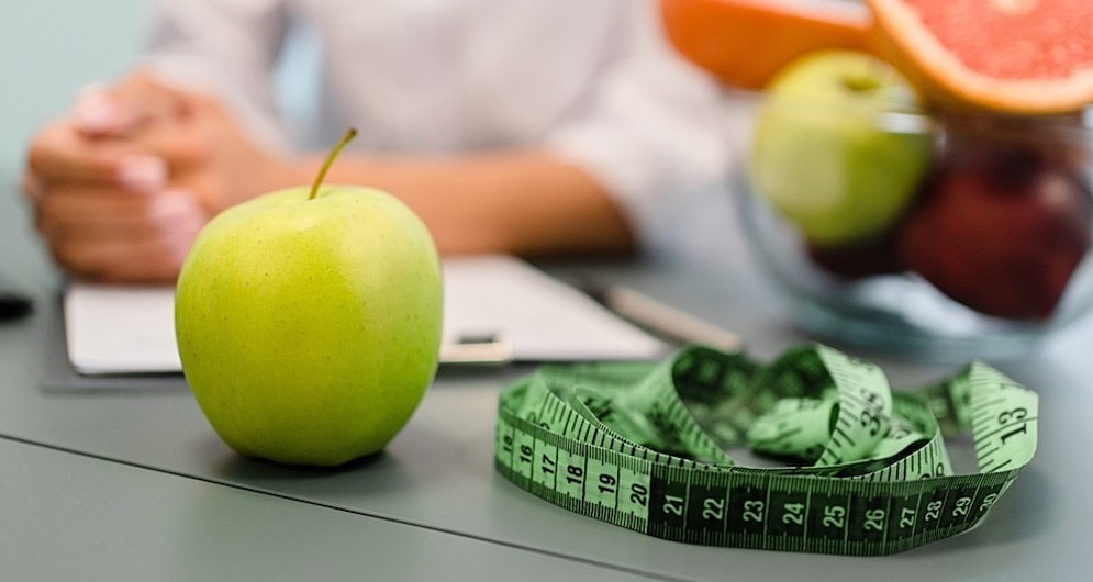 A bowl of fruit and a measuring tape on a desk, with a person’s arms and torso out of focus in the background
