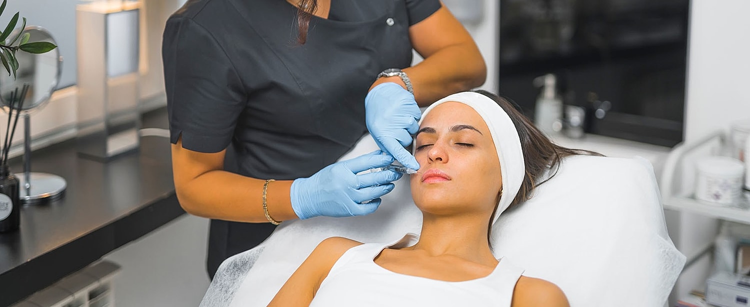 A practitioner in blue gloves injects a syringe into a woman’s upper lip as she lies on a treatment table with eyes closed.