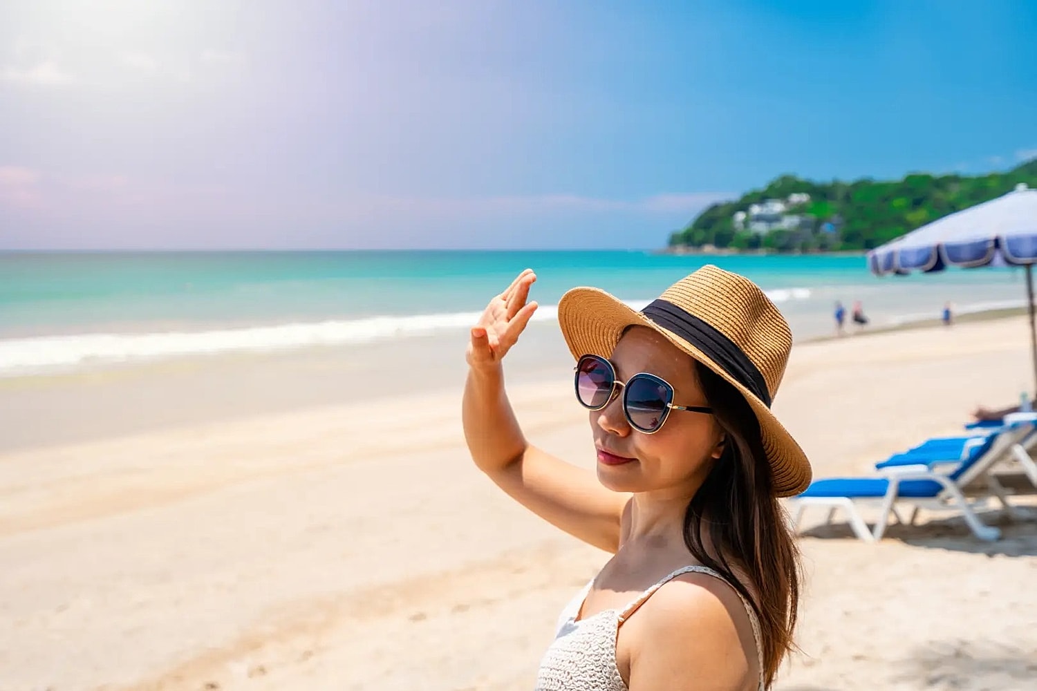 Woman wearing sunglasses and a straw hat raising her hand while standing on a sunny beach.