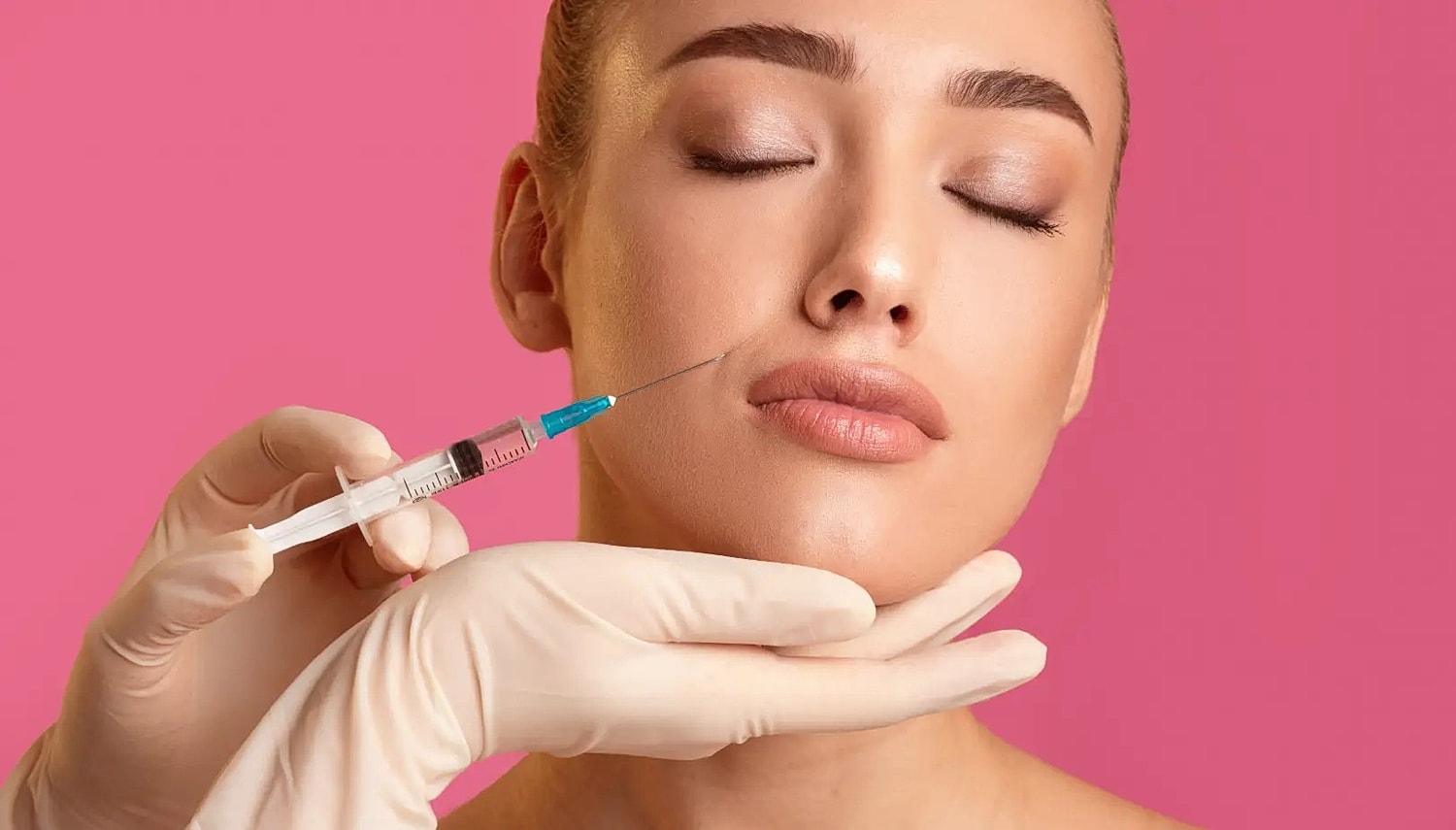 Gloved hands holding a syringe near a woman’s cheek against a pink background