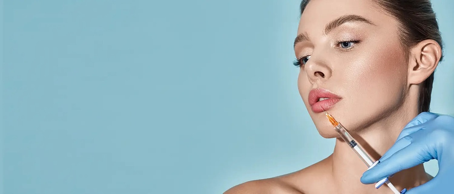 Gloved hand holding a syringe near a woman’s lips against a blue background