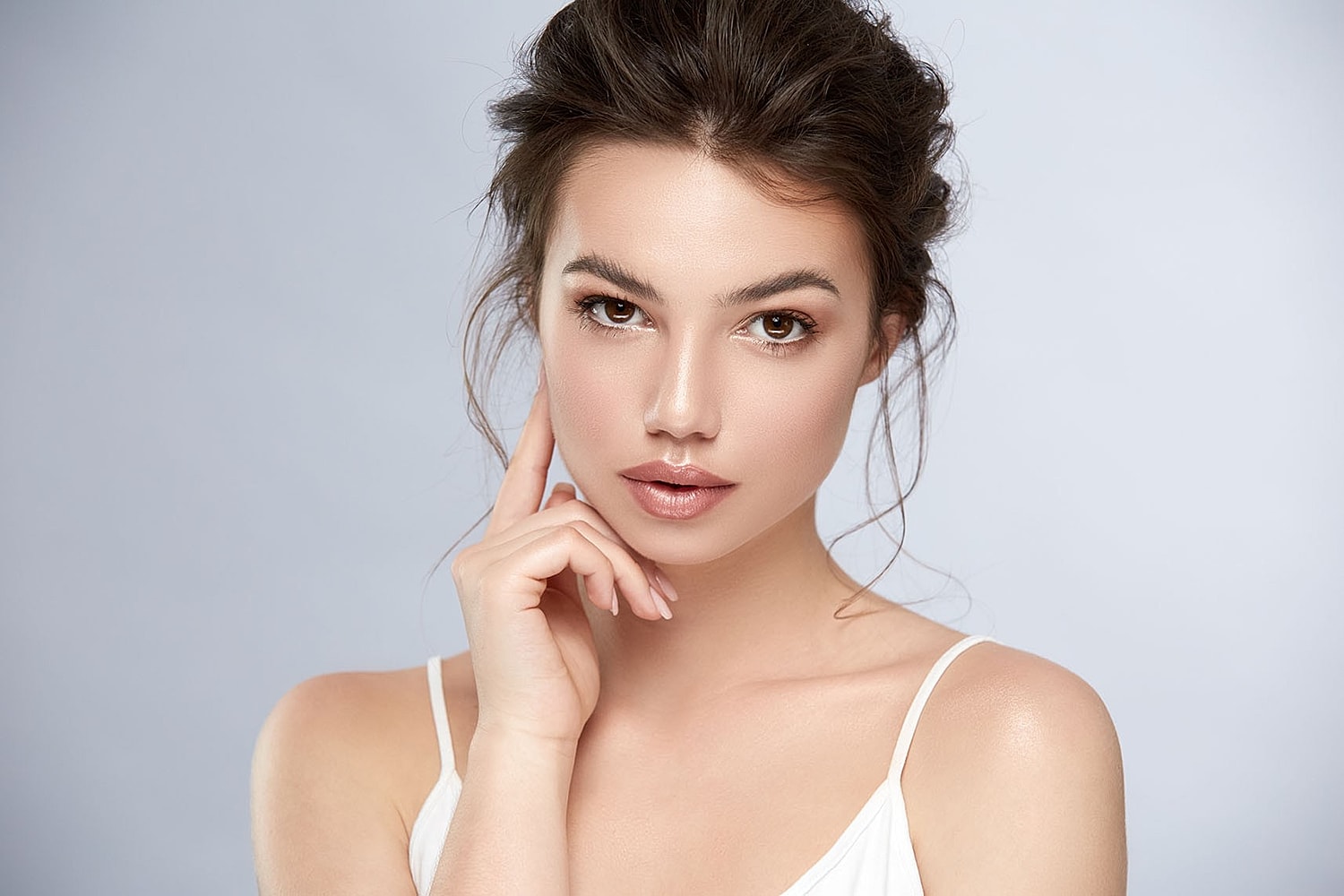 A woman looks toward the camera with one hand resting lightly on her cheek against a soft, light background.