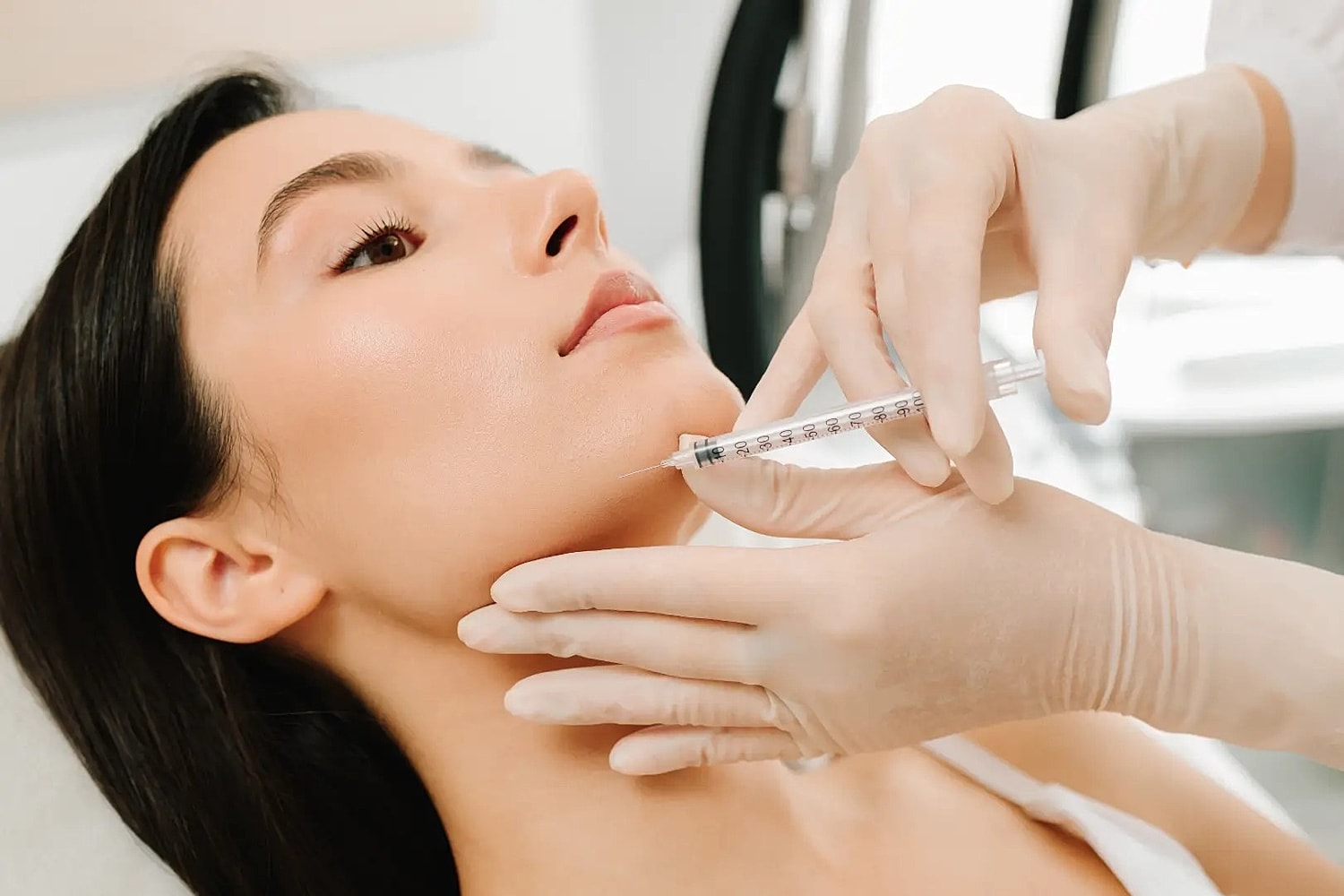 Gloved practitioner holding a syringe near a woman's lower jaw while supporting her chin.