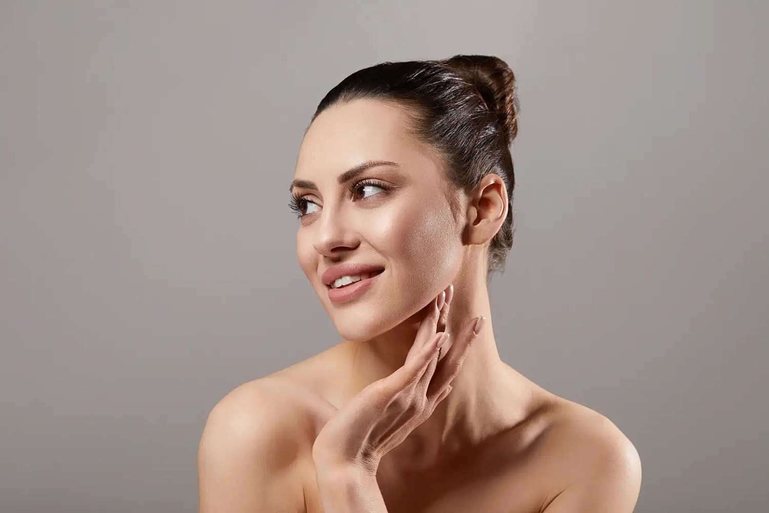 Woman with her hair in a bun touching the side of her jaw and neck against a gray background.