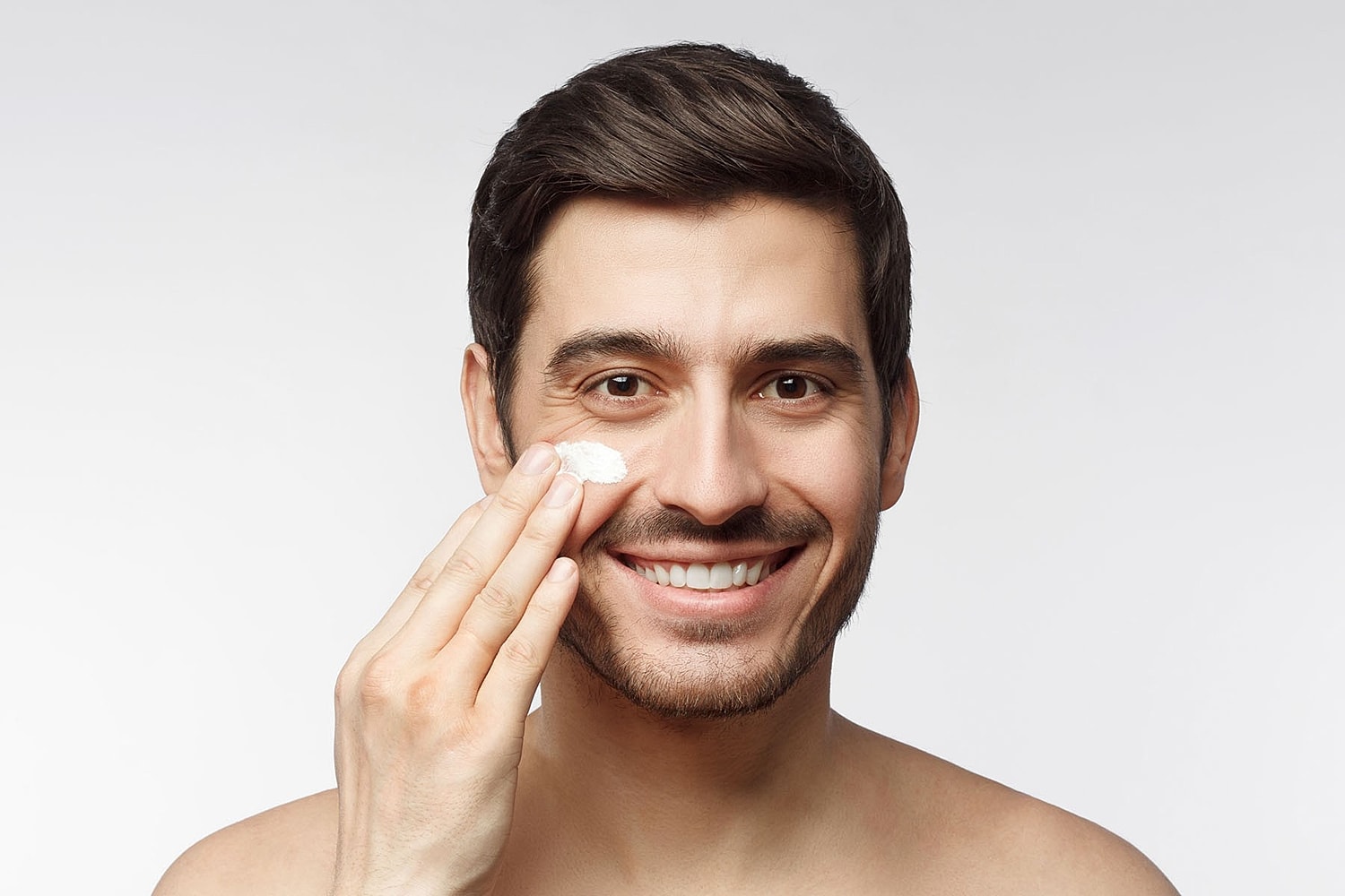 A man smiles while applying a small amount of cream to his cheek with his fingers.