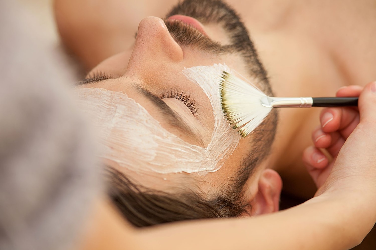 A man lies with eyes closed as a brush applies a white mask across his face.