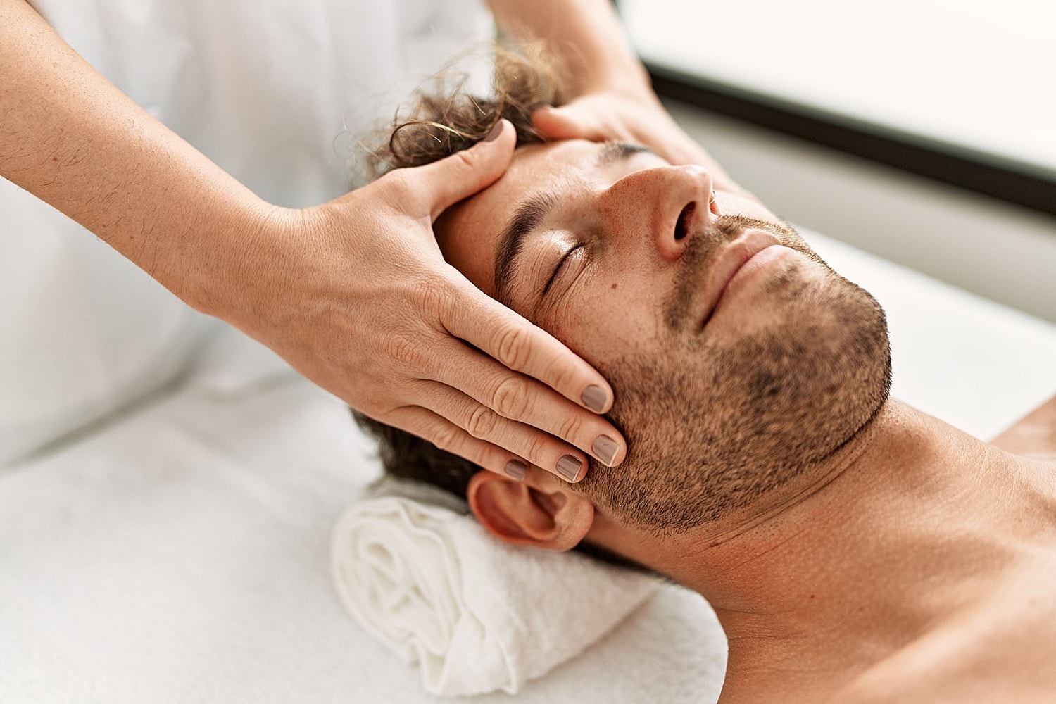 A man lies on a treatment table with eyes closed while hands gently hold the sides of his face.