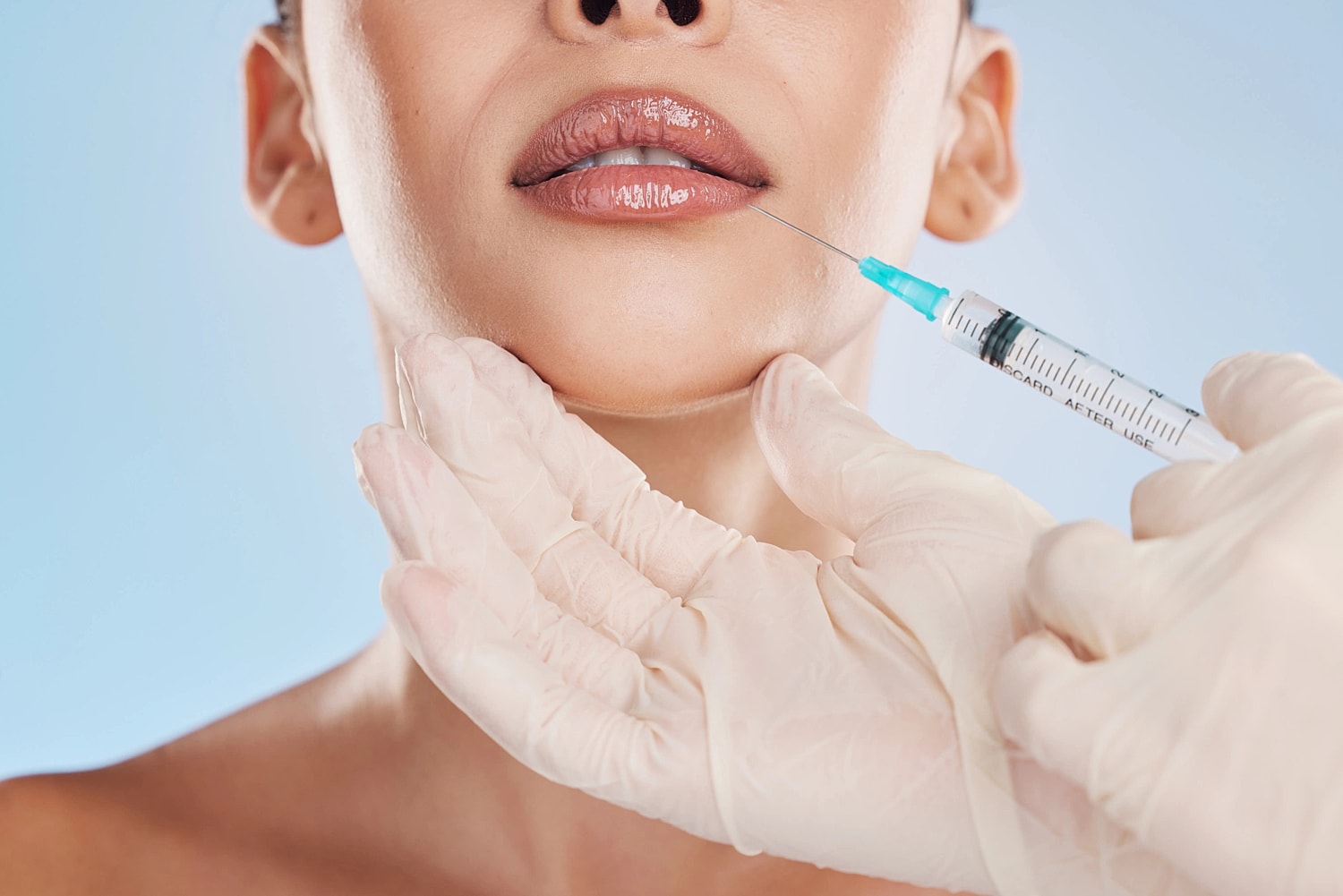 Close-up of glossy lips with gloved hands supporting the chin and a syringe positioned near the lower lip.