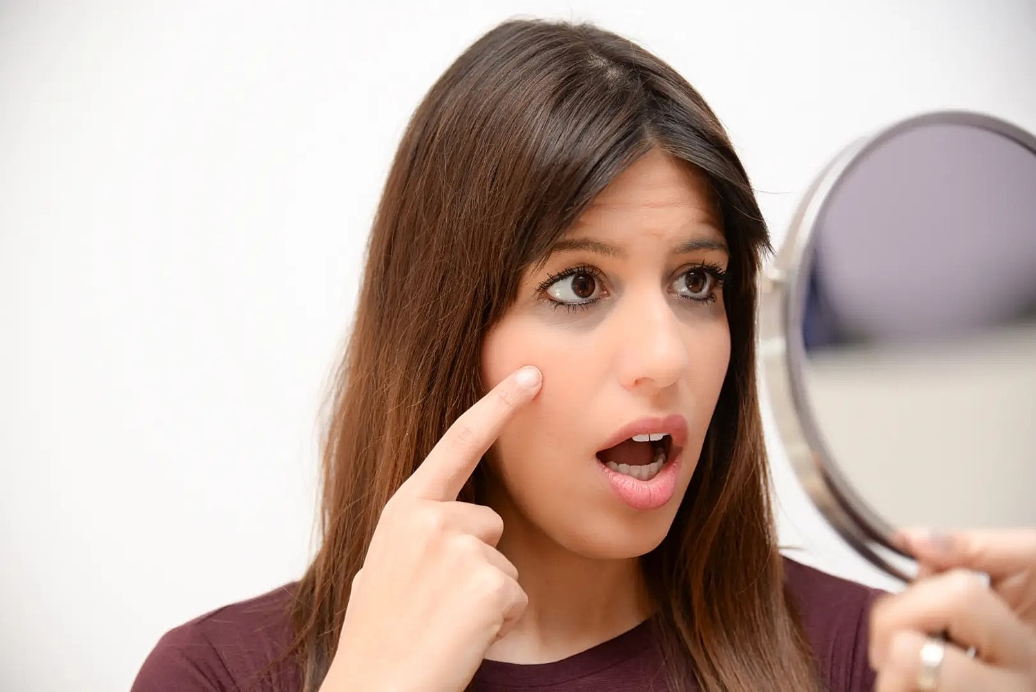 Woman looks into a handheld mirror while pointing to the skin beneath one eye.