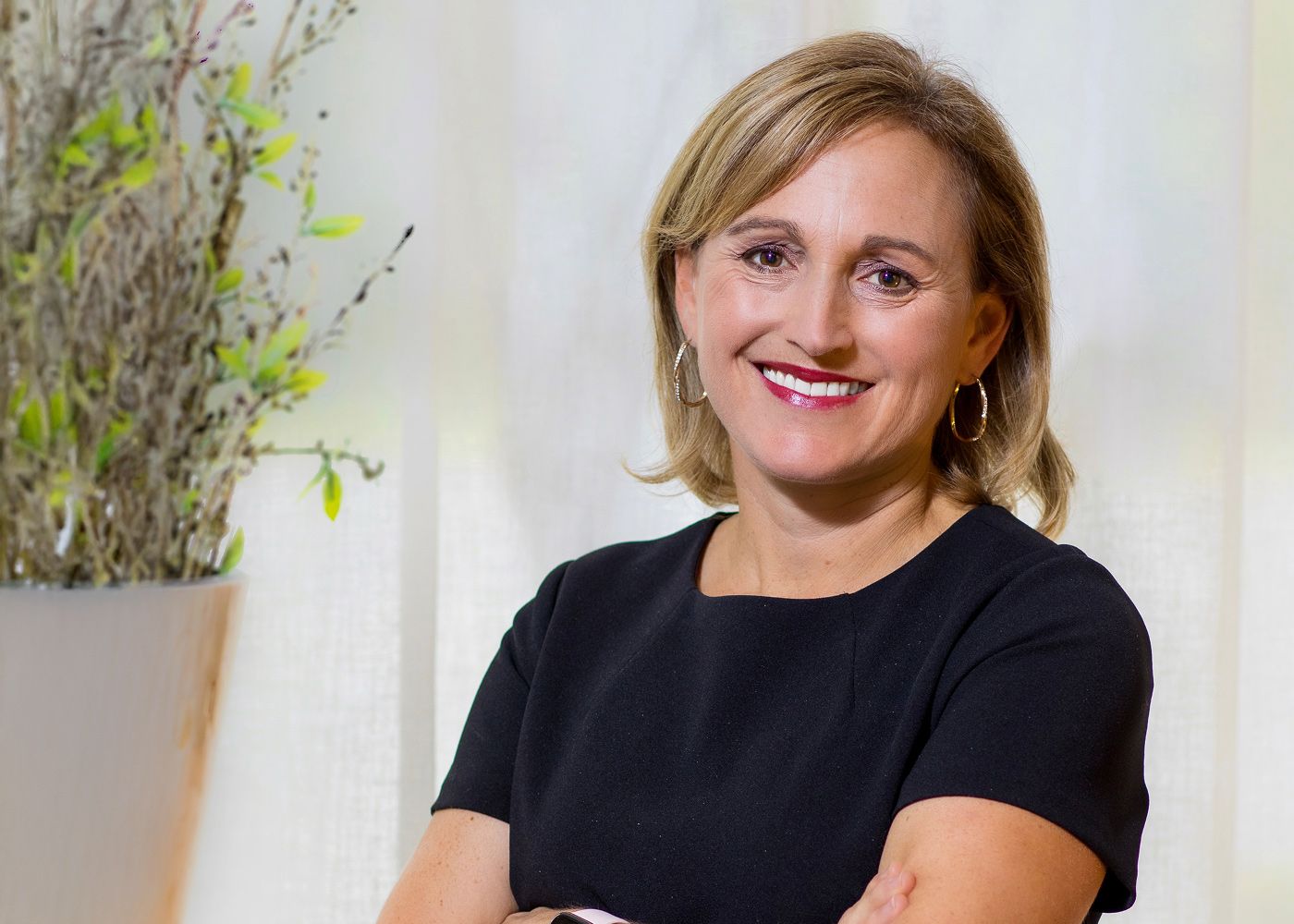 Smiling woman in black dress with plant backdrop.