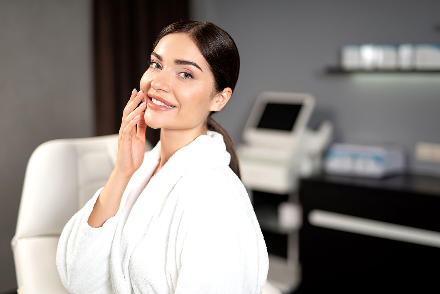 Woman in a robe smiling in a facial plastic surgery consultation room, representing facial rejuvenation procedures such as facelift, brow lift, blepharoplasty, and neck lift.