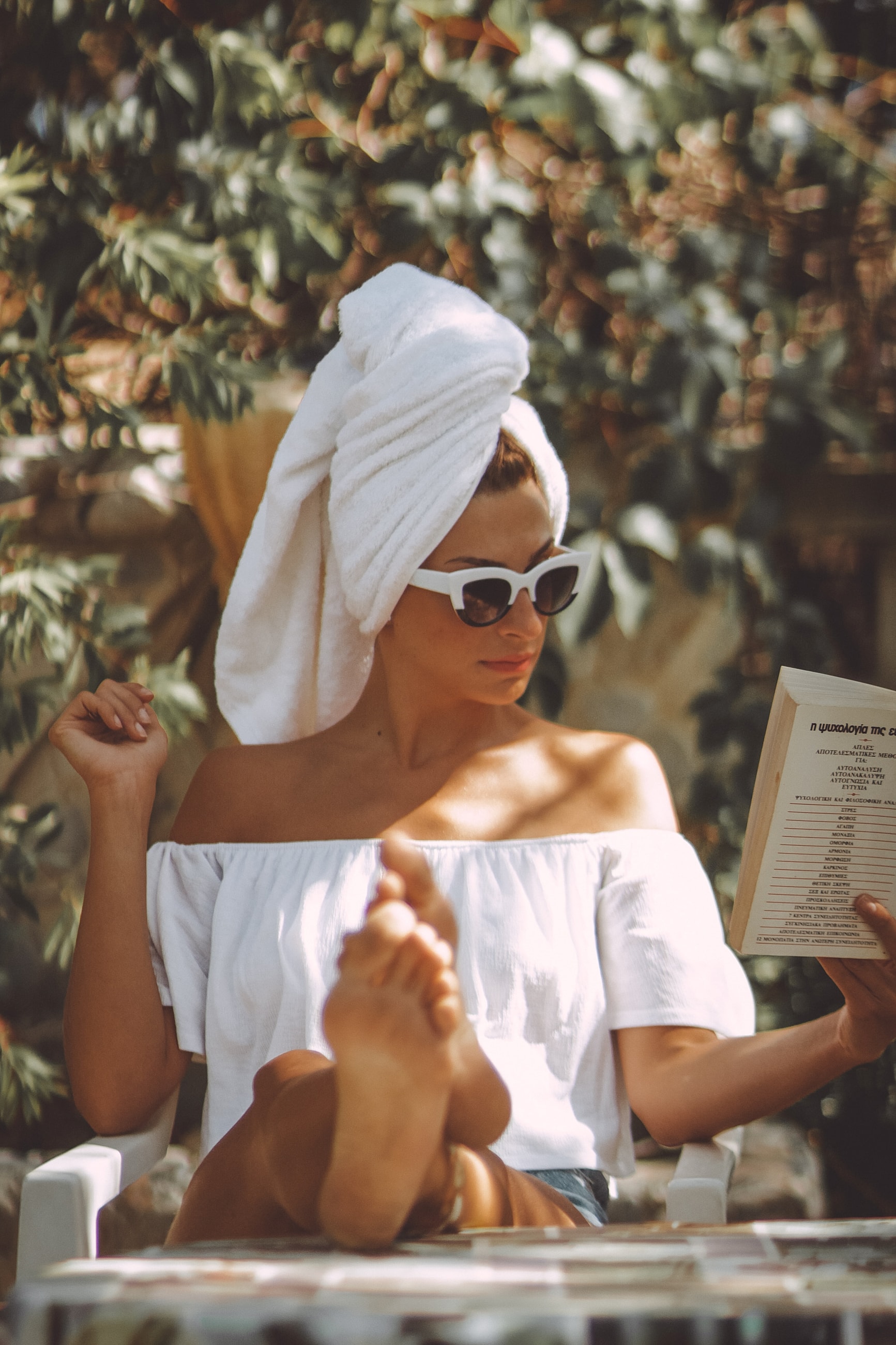 A woman enjoying a quiet, sunlit moment with a book, wearing sunglasses and a towel wrapped around her hair.