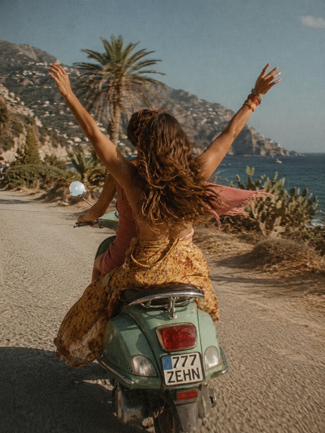 A woman raising her arms while riding as a passenger on a seafoam Vespa along the coast.